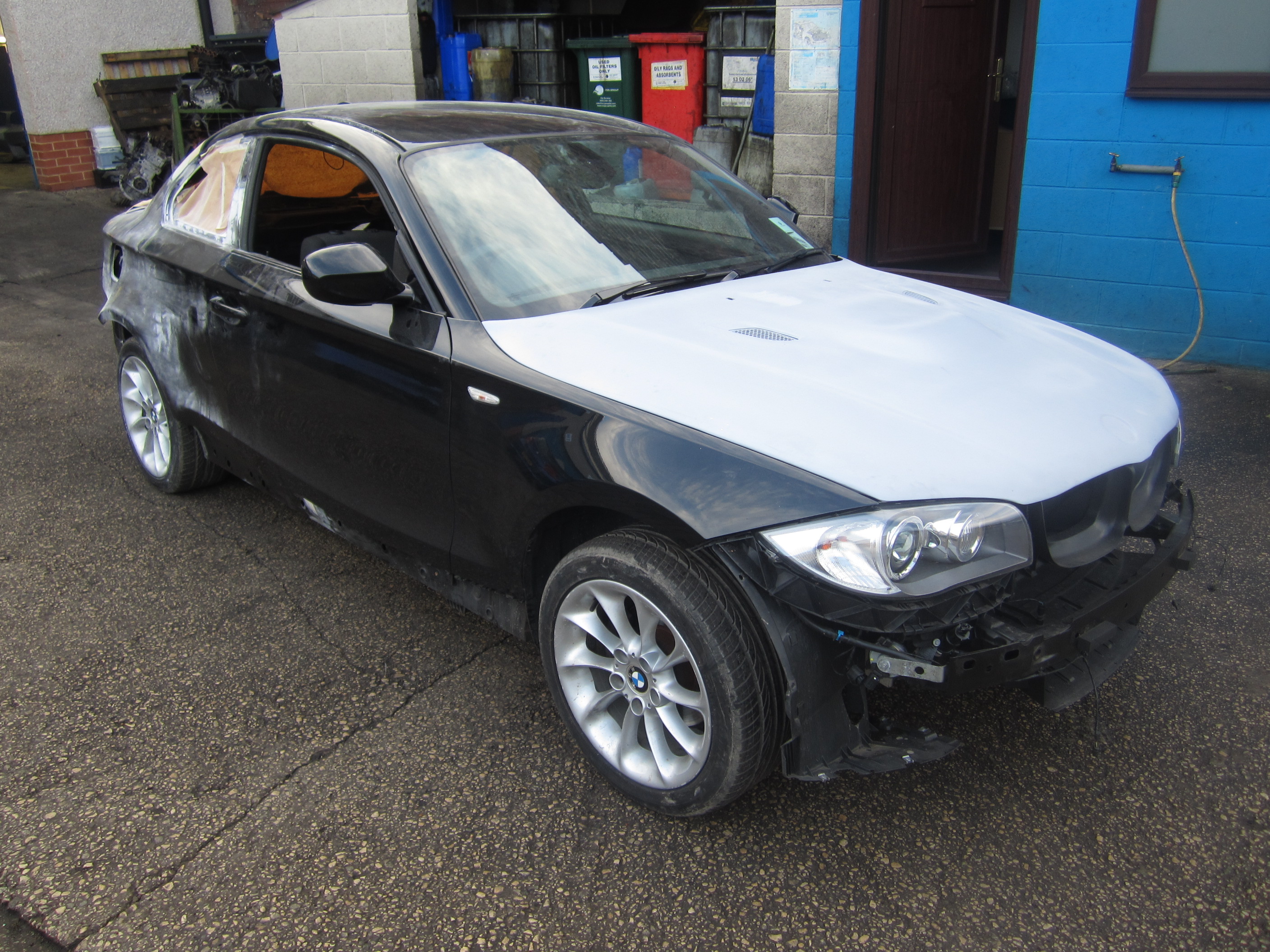 BMW 1M 4.0 V8 M3 Powered - Page 1 - BMW General - PistonHeads - This is a color photograph showing the front end of a compact car. It appears to be a BMW model, given the distinctive kidney grille and headlights. The hood is visibly damaged with scratches and dents, indicating the vehicle has been involved in some sort of accident. The car is parked in a driveway or yard, next to a blue wall. There's a large area of body panel missing or damaged, exposing the structure beneath. The surroundings include a red mailbox and a white building with a blue floor.