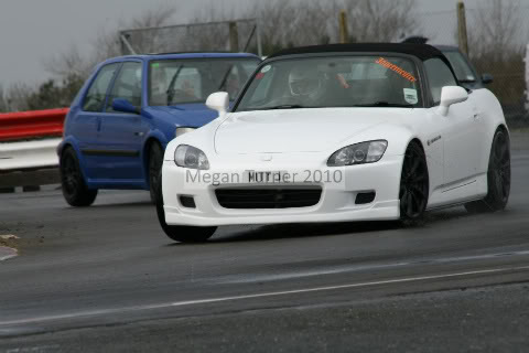 Pistonheads - The image shows a white sports car in motion on a racetrack, with another blue car in the background. The white car is the focal point of the photo, appearing sleek and shiny in the sunlight. The racetrack appears to be dry and well-maintained, with clear lane markings. The photo is taken from the perspective inside another vehicle, possibly capturing the moment the driver of the white car was in front of the photographer's car.