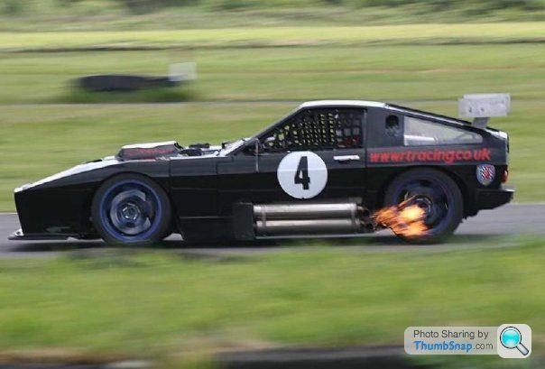 Thorpedo is back! - Page 1 - Sports - PistonHeads - The image captures a dramatic scene of a steel track race. The car, dominant in the frame, is a black vehicle adorned with a large white number four on the side. The race car is leaving a fiery trail behind, suggesting it's moving at a high speed. There's a metallic construction crane in the background, although it isn't clear if this adds to the context of the race. The background also features a grassy expanse with trees, which provides a calm contrast to the intense action in the foreground.