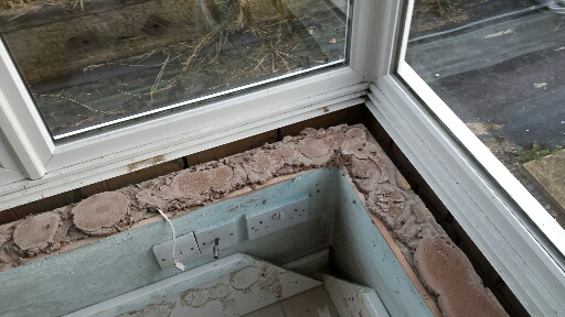 Damp walls in conservatory - suggestions? - Page 1 - Homes, Gardens and DIY - PistonHeads - The image shows a view from a window overlooking what appears to be a bathroom renovation in progress. The vantage point from the lower left side of the window provides a diagonal angle of the bathroom space. From the window, you can see exposed floorboards and a partial view of the room. Behind the window, there are branches, suggesting a garden or outdoor view. The room's concrete floor has areas missing, suggesting that the bathroom is under repair.