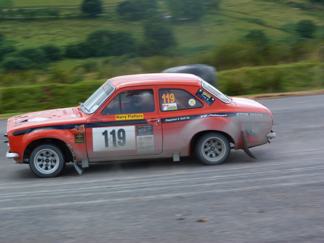 Why are there so few car photographs? - Page 117 - Photography & Video - PistonHeads - The image shows a vintage red car with white numerals, caught in mid-motion against a blurred greenish-gray backdrop that suggests speed. The car's paintwork appears to be aged, and it has a persistent, flowing mist around it that enhances the sense of motion. Its wheels and engine also bear the number 119, contributing to its dynamic activity. The car is being driven around a bend, indicated by the course it takes on a paved road, framed against a flowing field in the background. The style of the car suggests it is a race or rally car, referenced by the number and the aggressive driving posture.