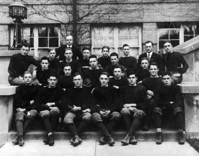 This is a black and white photograph showing a group of men posing together. They are dressed in formal attire, which suggests they are dressed for a formal occasion. The seated men are neatly arranged on at least two levels of a stepped area, and some are also standing around them. There is a building with visible windows in the background, and the men are positioned in such a way that they appear to be in a room with one wall visible. The composition and attire of the men suggest this may be an old photograph, possibly documenting a historical event or team.