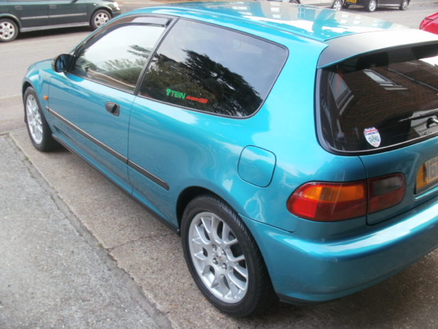 A car is parked on the side of the road - Pistonheads - The image showcases a vibrant turquoise car parked in a lot. The frame captures a partial view of the car, focusing on its side profile that highlights the modern design and glossy finish. The back door is shut, indicating that the car is parked securely. In the background, several other cars are visible, parked in a neat row, creating a typical parking scene. The angled view of the image and the car's color make it a dynamic and visually appealing image.