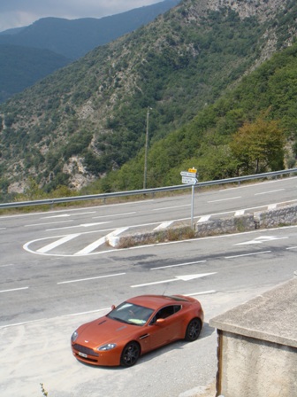 Week Pistonheads Turini - The image depicts a vibrant scene on a Highway in the mountains. An orange car, sleek and shiny, is prominently parked on the shoulder of the road. The car's bright color contrasts beautifully with the lush green of the mountains and the hazy blue of the sky. In the background, a signpost can be seen, standing out against the backdrop of the highway. The relative position of the objects suggests a peaceful day with no immediate danger or traffic. Overall, the image captures a moment of stillness amidst the anticipation of a journey.