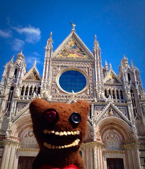 Pistonheads - The image features a statute of a large, brown bear character with visible teeth and a peeling, moist appearance standing in front of a grand, medieval-style cathedral. The cathedral has multiple steeples and intricate details, and the sky is clear with a few scattered clouds. The bear character appears to be posing for the photo with its mouth open as if it's about to say or eat something, which creates a humorous and unexpected contrast with the reverential presence of the cathedral.