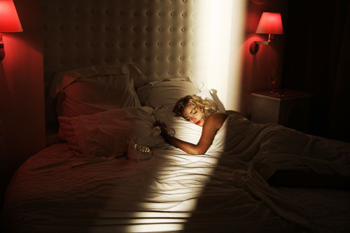 The image is a photograph of a dimly lit room with a superimposed figure. The central figure appears to be a woman lying on her side in bed, facing away from the camera. She is depicted with her eyes closed and posture indicating she is asleep or resting. The room is softly illuminated, possibly by the glow of the bed lamps with red shades, creating a cozy and intimate atmosphere. The bed has a white comforter and there is at least one pillow visible. The photo exhibits a warm, muted color tone, primarily focusing on dark, red-hued lighting.