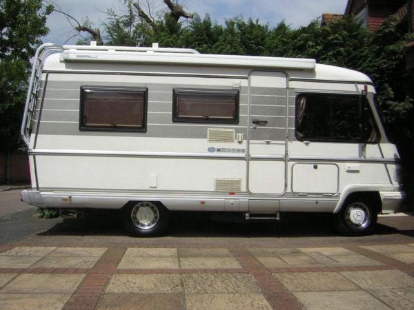 Campers / motorhomes short term ownership - Page 1 - Tents, Caravans & Motorhomes - PistonHeads - The image showcases a white travel trailer parked on a driveway in front of a house. The trailer is rectangular with two windows on the side, and a camper door at the back. It has a bumper, towing this detachable structure. The driveway is visible, leading to what appears to be a residential street. The trailer sits atop a patio surrounded by a concrete path, with a verdant tree providing a refreshing backdrop. The trailer's position and the overall setting suggest a sense of tranquility and independence.