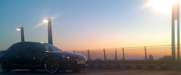 Pistonheads - The image shows a black Mazda Miata convertible car parked on the side of a road during sunset. The vehicle's top is down, and its shiny exterior reflects the day's last light with the golden hues of the setting sun in the background. The scene is fenced in by a metal railing, indicating this might be a designated parking or viewing area. There are tall lights perpendicular to the rods, casting a glow that contrasts with the sky's colors.