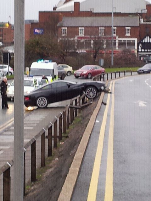 Vanquish Crash! - Page 1 - Aston Martin - PistonHeads - The image shows a significant road accident in which two cars have collided and are tipped over, on or near a divider. Emergency services are at the scene, with police cars and ambulances present, as well as personnel tending to the incident. The road is blocked, and the emergency response indicates a major disruption. There are no visible signs of individuals in distress or the intensity of the crash.