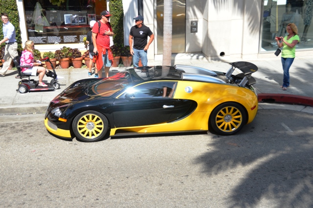 Supercars spotted, some rarities (vol 6) - Page 35 - General Gassing - PistonHeads - The image captures a vibrant street scene centered around a bright yellow and black Bugatti sports car. The Bugatti, with its large side scoops and distinct wheels, is the primary focus of the image. It is parked in the middle of a curb, on what appears to be a sunny day.

Around the car, there is a lively atmosphere with several people walking on the sidewalk. One individual is seen pushing a stroller, while others are casually strolling, likely going about their day. A few potted plants add greenery to the urban setting, placed here and there along the curb.

Further adding to the urban vibe, a few shops are visible in the background. One is with a red awning, and another with a green one, both inviting passersby with their colorful entrances.

The street itself is bathed in sunlight, creating beautiful shadows on the ground and enhancing the overall warmth and vibrancy of the scene. The bright sunlight provides a pleasant contrast to the sleek, dark color of the Bugatti, making it stand out even more.