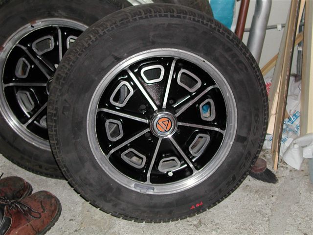 Ginetta Pistonheads Wanted Alloy Rim - The image shows two large vehicle tires resting on a floor. Both tires are black with silver rim hubs. One of the tires has a red and yellow sticker on the center of the hubcap. The tires appear to be used and are not on a vehicle. The background is slightly blurred, drawing focus to the tires, but includes a hint of mechanical or building parts.