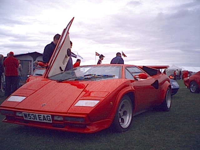 Pistonheads - In the midst of a bustling event, a striking red Ferrari sports car commands attention. The physical condition of the car is pristine, indicating careful maintenance. Its top is down, exposing a vibrant red interior that matches the exterior. Surrounding the car, a crowd of people interact, some closer to the car while others are at a distance, all seeming to enjoy the scene. The background is filled with blurred figures and cars, adding to the lively atmosphere of the car show.