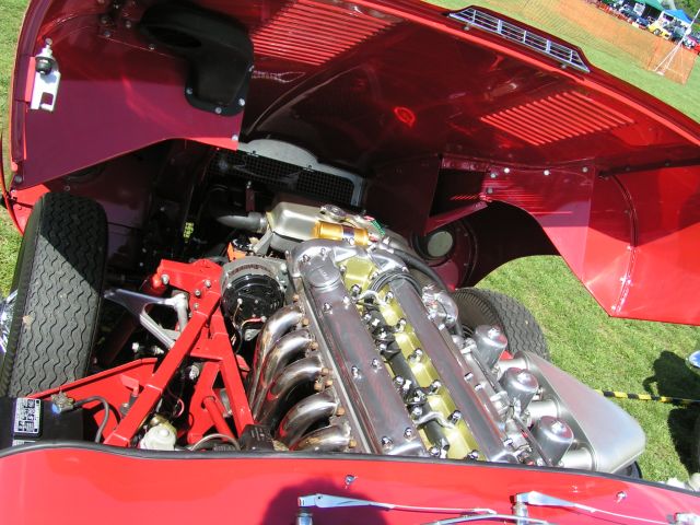 Pistonheads - The image showcases the front and undercarriage of a red race car. It has an open hood displaying its powerful engine, which appears to be a V-12, and numerous carburetors. The engine is set against the backdrop of track mesh fencing, suggesting the scene is at a racetrack or motorsport event. The red color of the car contrasts with the metallic sheen of the engine components, emphasizing the mechanical beauty of the vehicle.