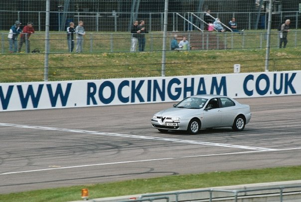 Pistonheads - The image shows a small, grey car with a license plate that reads "V5211X" on a racetrack. The car is driving past a large blue and white advertisement sign that reads "WWW, ROCKINGHAM, COUK, VW join us." The track itself is large, and there are several spectators visible in the stands, some of whom are seated and others standing. The spectators seem to be focused on the car, suggesting that it might be participating in a race or a demonstration event.