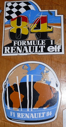 Show Us Your Stickers! - Page 1 - Formula 1 - PistonHeads - The image features a collection of stickers related to Formula 1. At the center, there's a large sticker with the words "Formule 1 Renault Elf" around a graphic of a simplified world map. Surrounding this central sticker are two smaller stickers. One has a large "84" prominently displayed, suggesting an association with the Renault 84, a Formula 1 car from the 1984 season. The other small sticker features black and white checkered patterns, a design often associated with racing cars, particularly Formula 1. The stickers are mounted on a wooden surface.