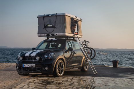 Small engined first car with fold flat rear seats for sleep? - Page 1 - General Gassing - PistonHeads - In the image, a black Mini Cooper car is loaded for a journey, specifically camping. The car is parked on a stone surface, facing towards the left side of the image. Attached to the trunk of the car is a rooftop bike carrier with two bicycles securely fastened. On top of the caravan, there's an enclosed sleeping area, providing a makeshift camper's refuge. The overall scene suggests an adventurous road trip with the car and bicycles ready to explore the destination's natural beauty.