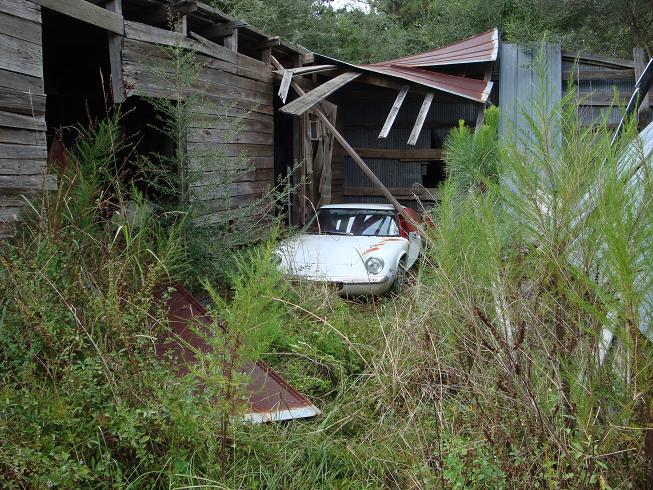 Tow point on S2 Europa? - Page 1 - General Lotus Stuff - PistonHeads - The image shows an old, white sports car that appears to be abandoned or in a state of disrepair. The car is situated in a surroundings covered with overgrowth, weeds, and tall grass, suggesting it has been sitting there for some time. The car is in a wooded area and near a dilapidated structure, which, along with the car, evokes a sense of abandonment and neglect. The car has a vintage design, featuring a long hood and short rear deck, typical of the sports car type.