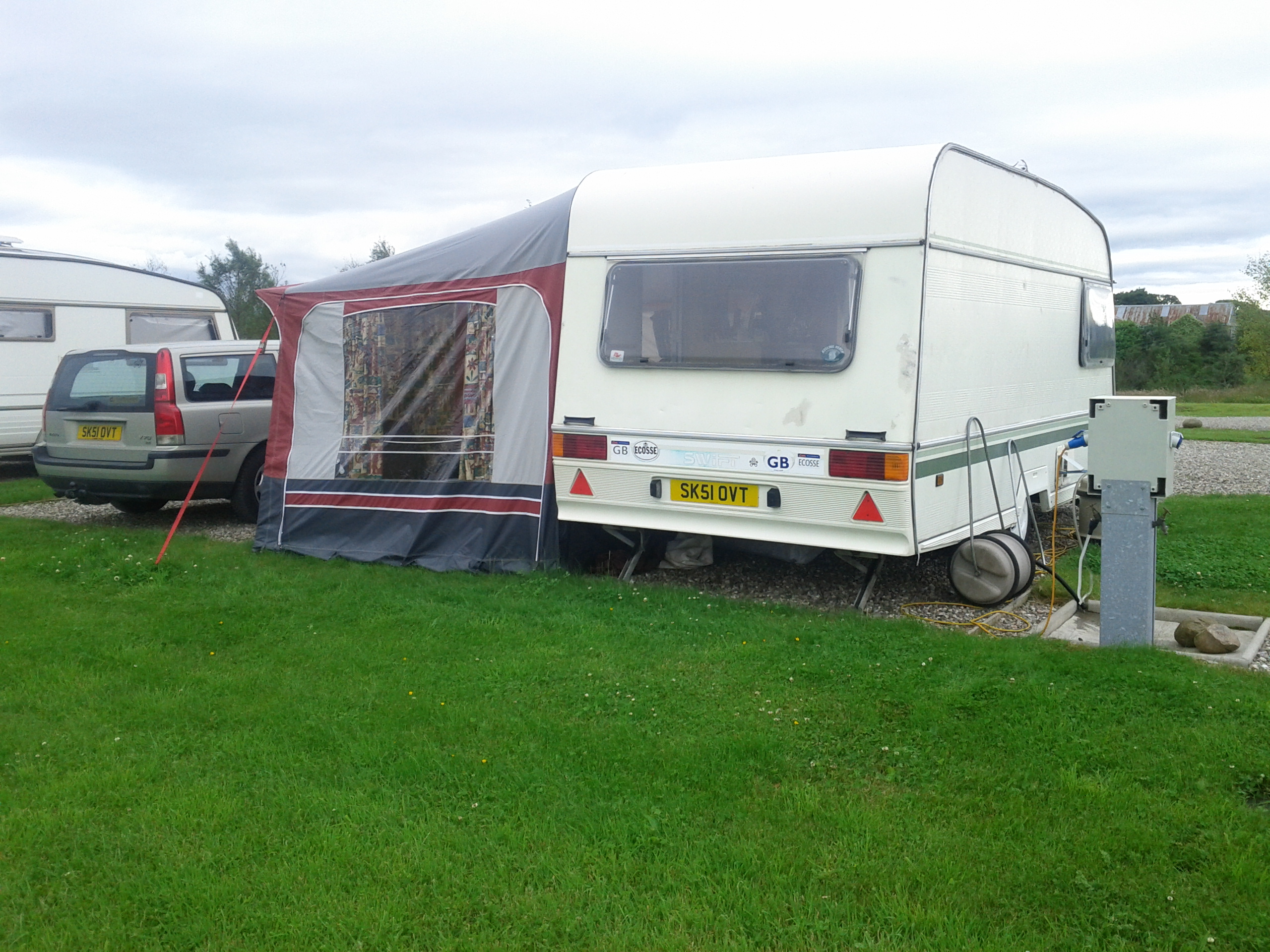 Useable caravan for £500? - Page 1 - Tents, Caravans & Motorhomes - PistonHeads - In the image, there are two trailers stationed on a grassy landscape. The larger one has a distinctive white and gray exterior with red stripes along the bottom. It features a ground-level door and a large rectangular window. Attached to it is a small one, which has a red awning. Both trailers have a European-style license plate on the front. The trailers are parked in a lot that is lined with larger vehicles, including a tall white caravan and a minivan. The sky appears overcast, suggesting it might be a cool or cloudy day.