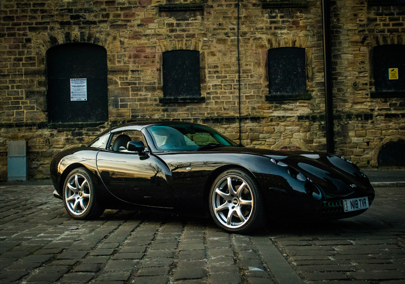 Free photoshoot opportunity  - Page 1 - Yorkshire - PistonHeads - The image displays a black convertible sports car parked on a cobblestone street in front of a brick building. The car is positioned at a slight angle, and the retractable top is down, revealing its interior. The windows of the building behind the car are closed, and the car's distinctive design features aerodynamic lines and a low-slung profile typical of sports cars. The lighting appears to be natural, suggesting it could be dawn or dusk. The image has a clear, high-resolution, providing a detailed view of the car's design and the ambiance of the street.