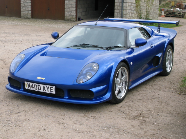 A blue car parked in a parking lot - Pistonheads - The image showcases a blue sports car parked on a dirt surface. The car's design is sleek and modern, with a low front profile. It features a prominent rear wing, a unique design element that adds to its aerodynamic appearance. The vehicle is positioned in the center of the frame, making it the main focus of the image. The surrounding area is mostly barren, with a few wooden structures visible in the background. There are no visible texts or distinctive marks to provide additional context about the car or its location.