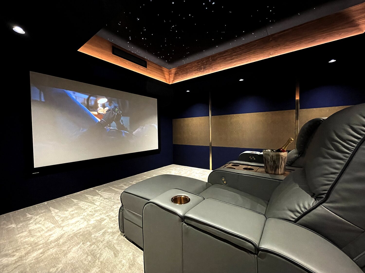 Cinema rooms - what have you got? - Page 12 - Home Cinema & Hi-Fi - PistonHeads UK - The image shows a modern and luxurious home theater room. In the foreground, there is a comfortable black leather recliner with cup holders, inviting viewers for an immersive experience. To the right of this chair, there's a large screen displaying a movie, suggesting that the room is in use.

The walls of the theater are adorned with dark blue acoustic panels to enhance audio quality. The floor appears to be carpeted, and the overall color scheme includes shades of blue, grey, and brown, creating a cohesive and sophisticated aesthetic.

On the left side of the room, there's a projector screen that is currently not in use, hinting at the possibility of alternative media consumption. The ceiling features recessed lighting, contributing to the overall ambiance of the space. There are no visible texts or distinctive brands within the image, and the focus is clearly on the home theater setup.