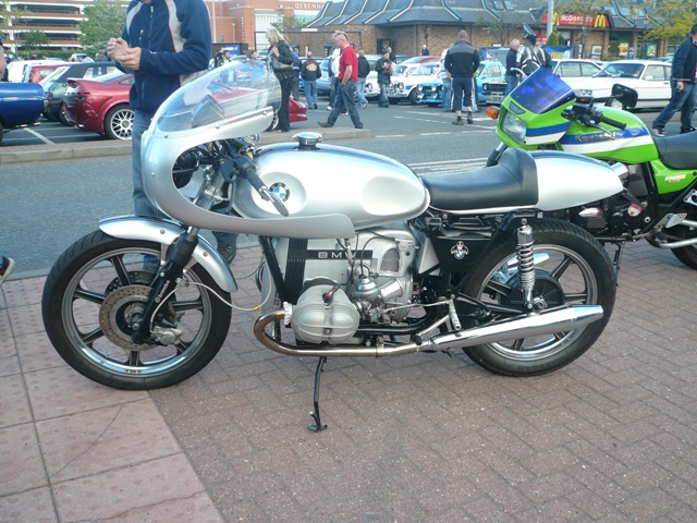 Pistonheads Metrocentre - The image shows a collection of motorcycles parked on a paved area, possibly a parking lot or a street curb. The main focus is a unique BMW motorcycle with a prominent headlight, and it is surrounded by various other motorcycles. Some of these motorcycles have distinctive colors and designs, suggesting they might be custom or special edition models. The crowd in the background, along with the diverse assortment of vehicles including cars and trucks, indicates a public event or gathering might be taking place.