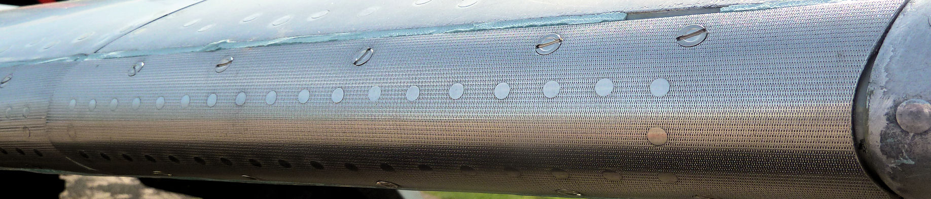 April Model Raf Cosford Pistonheads - The image shows a close-up view of a metallic surface, likely the side of an engine or tank, with a design that resembles a wire mesh pattern. The surface is reflective and has a series of holes or perforations which are possibly made for ventilation or structural support. There are water droplets on the surface, indicating that the photo may have been taken during or after a rain. The background is out of focus, emphasizing the texture and pattern on the metallic surface.