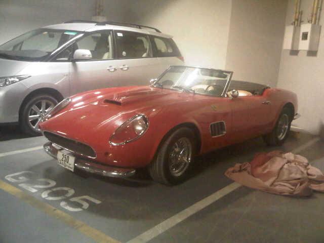 A car with a surfboard strapped to the back - Pistonheads - In the image, a sleek red sports car is the main focus, parked in an indoor parking lot. The car's shiny exterior reflects the ambient light, and it is situated next to a white van. Additionally, a dirt-covered cover can be seen in the foreground and some pipes are visible above the car. The parking lot has a gray floor and few other vehicles can be seen in the background. The overall scene suggests a garage-like atmosphere where the sports car is perhaps being stored or maintained.