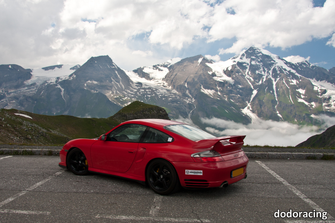 A car parked on the side of the road - Pistonheads - The image features a vibrant red Porsche nine generation sports car parked prominently in the foreground. Behind it stands a breathtaking mountainous backdrop, heavily blanketed in snow on the meringe-looking peaks. The contrast between the car's bright color and the glistening white snow creates a visually striking scene. The car is positioned on a clear, cloudless day, adding to the overall serenity of the landscape.