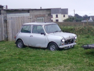 A white truck parked in a parking lot - Pistonheads - The image shows a small silver car parked on grass in a field. The car is facing forwards to the left side of the frame. The car is an older model, and its color is a uniform silver. The field is partially bordered by a weathered wooden fence on the left. In the distance, there's a building with a white exterior. The setting appears to be a rural or semi-rural area based on the vehicles' surroundings. The sky is overcast, suggesting an overcast or possibly rainy day.