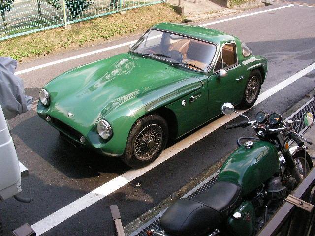 Early TVR Pictures - Page 67 - Classics - PistonHeads - The image displays a trio of vintage vehicles on what appears to be a roadway or street. Dominating the foreground is a green convertible car, its hood gleaming under the sunlight. Beside it, a sleek black motorcycle adds a contrasting element to the scene. The motorcycle is parked upright, its handlebars facing outwards, as if ready for departure at any moment. In the background, partially obscured by the car, is another vehicle that has been styled to look like a motorcycle, complete with the characteristic silhouette. This imaginative design plays with the viewer's perception of vehicles, challenging our expectations with an unexpected twist. The presence of these vehicles suggests a journey or adventure, possibly paused for an encounter at a grill event. Overall, the image captures a moment of stillness on a road where the old meets the new and imagination takes center stage.