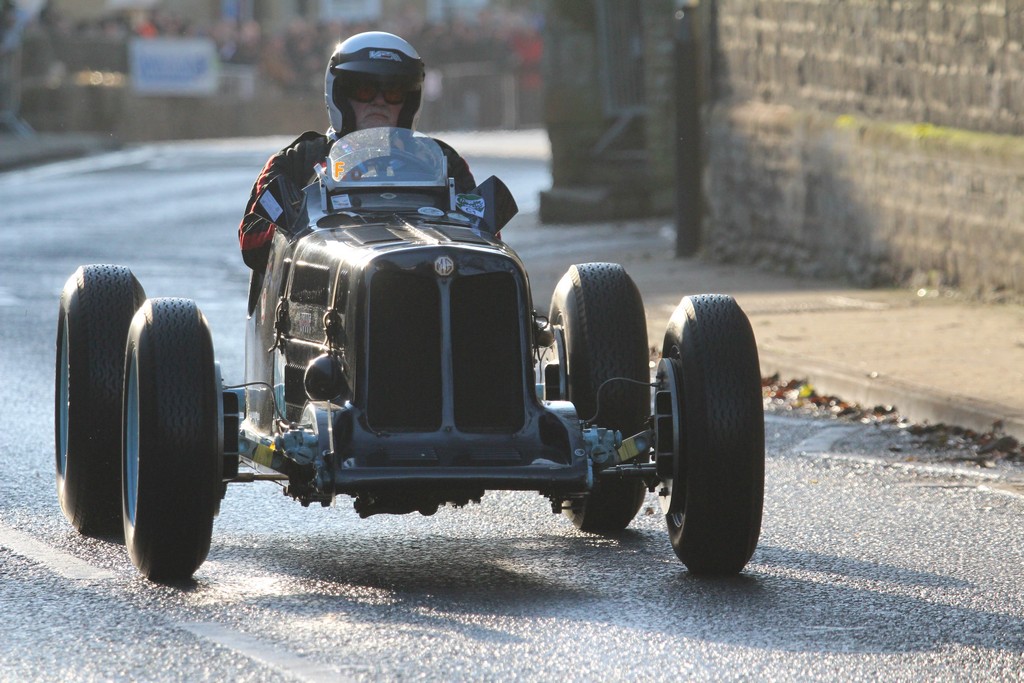 A man riding a motorcycle down a street - Pistonheads - The image captures a dynamic scene of a man racing a black and white 3-wheeled car down a wet roadway. The man, clad in a white helmet and goggles, is seated in the middle of the buggy-style vehicle. The vehicle's design, accented with a chrome grille and a single headlight on the front, lends it a vintage feel. The contrast between the white of the car and the black of the tires adds a striking visual element to the scene.