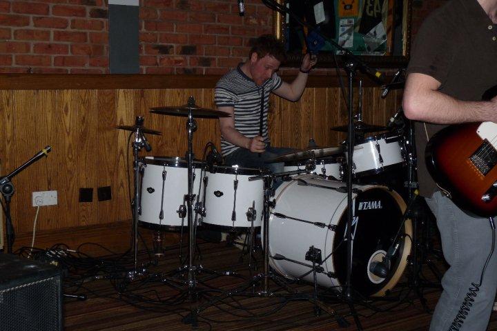 Pistonheads Musicians - The image shows an indoor music practice setting. A man is in the foreground sitting on a floor next to a white drum set. He appears to be interacting with the drums or waiting for his turn to play. To his right, there is another person with a guitar, suggesting a coordinated musical performance. The background features a brick wall, possibly a part of the venue, and a wooden panel, giving the setting a rustic vibe. The atmosphere seems casual and focused, typical of a musical practice session.