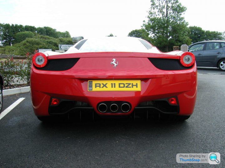 Breakfast Meeting, The Hare, Roxwell 4th June - Page 1 - Kent & Essex - PistonHeads - The image depicts a red Ferrari R8ruppe parked on a street. The car's license plate reads "RX11 DZH," and it features the distinctive emblem and design of the luxury sports car brand. The Ferrari is parked near a gray car. In the background, there is a line of trees and a sign indicating "Princess Risborough." The setting appears to be an urban or suburban location.