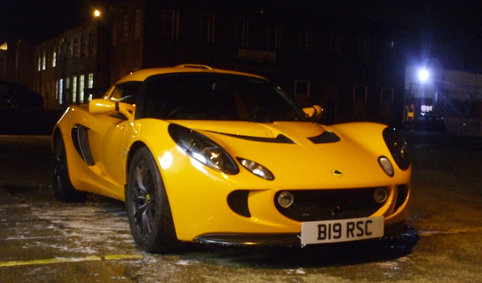 Pistonheads Exige Mini - The image features a bright yellow Mazda sports car parked on a dark street. The car is structured with a large front grille, two black wheels, and sleek body lines that suggest speed and performance. The illuminated headlights and the distinct badge on the grille are clear markers of the car brand. The street itself has scarce lighting, adding to the dramatic contrast of the car against its surroundings.