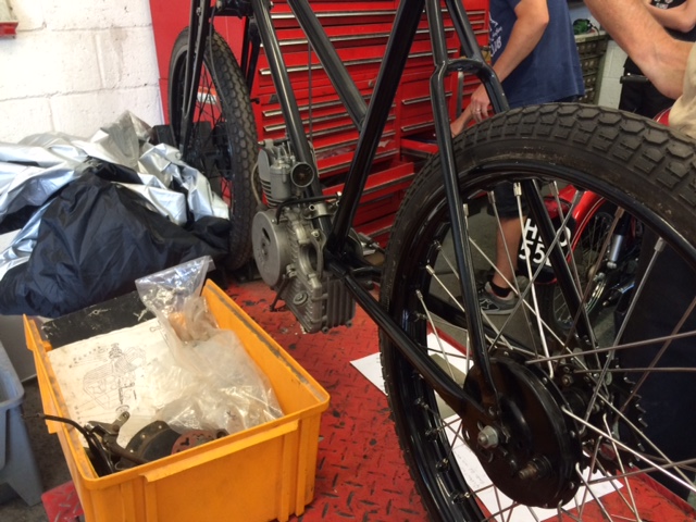 A picture a day....biker banter (Vol 4) - Page 505 - Biker Banter - PistonHeads - The image shows a bicycle partially disassembled in a workspace with tools on a table to the left. The bicycle's front wheel is prominently visible in the center of the image, and a red workbench framework can be seen in the background. A person, partially visible, is standing near an open red drawer to the left, suggesting that they might be working on this bicycle or another one. The bike appears to be in the process of being maintained or repaired due to the accessory attached to the handlebars.