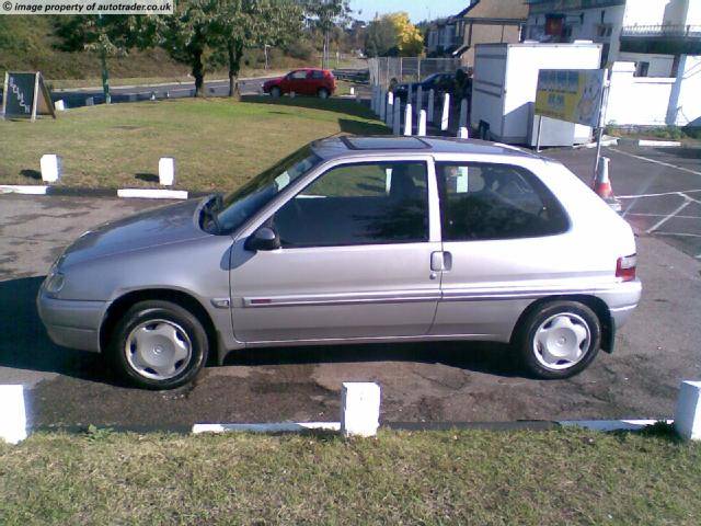 Saxo Pistonheads Citroen - The image features a gray, two-door hatchback parked in a space against a digital parking meter. The car has a passkey on the windshield. In the background, there are signs for parking rules, a lush green lawn with some bare patches, and a few vehicles in the distance, including a red truck. The scene appears to be in an outdoor parking area near a building or near a roadside.