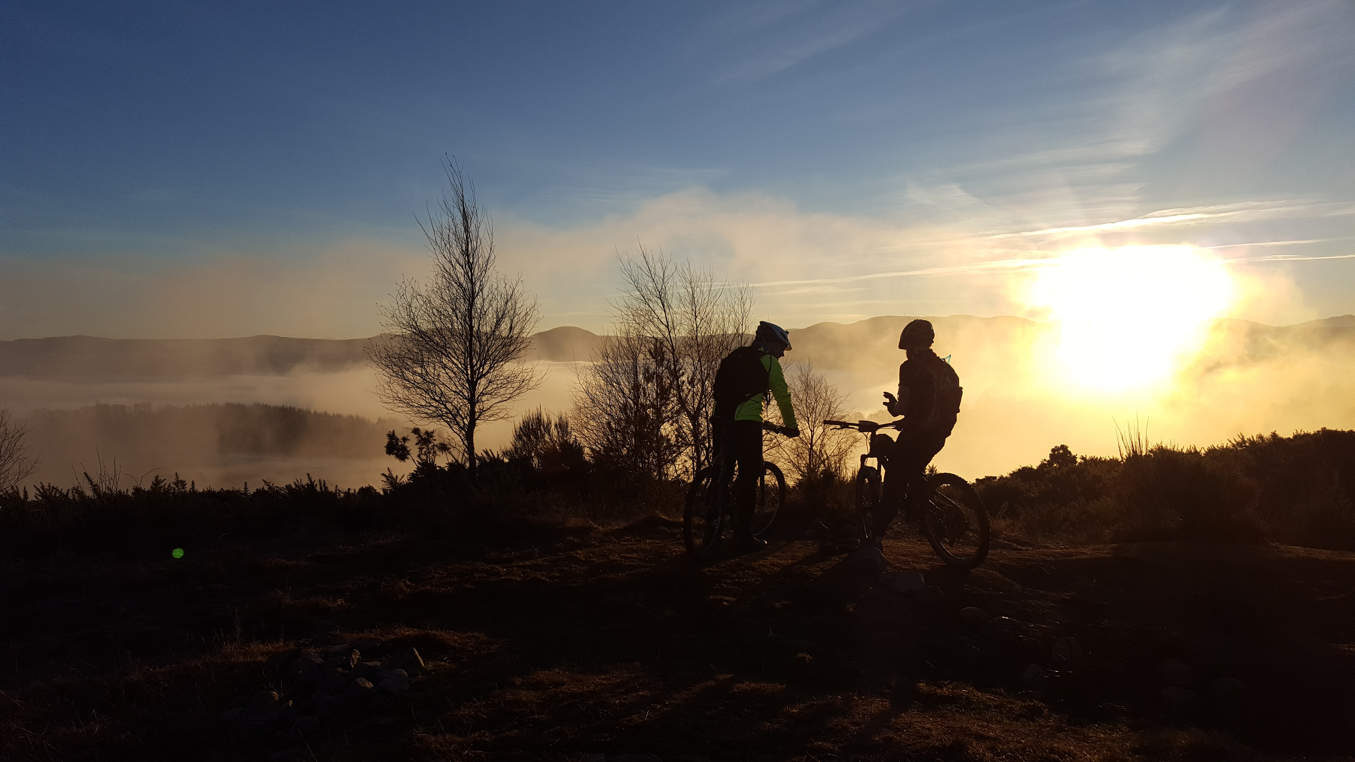 Random Photos : Part 4 - Page 357 - Photography & Video - PistonHeads - The image showcases two individuals engaged in a recreational activity, possibly mountain biking. They are standing on a dirt surface, with a bicycle prominently featured in the foreground. Both individuals are dressed in full-body apparel, suggesting a cool or windy climate. In the background, fog is resulting in a somewhat ethereal landscape, providing a sense of depth to the scene. The sun is setting, casting an orange glow on the right side of the photograph, highlighting the silhouettes of the cyclists and their surroundings.