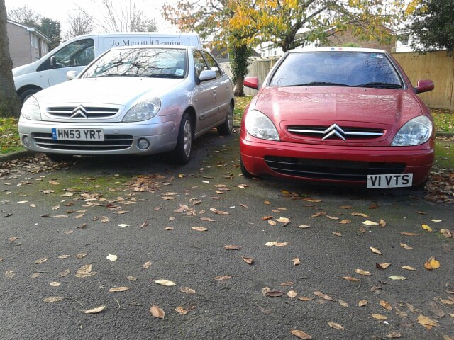 My Two French beauties. - Page 2 - French Bred - PistonHeads - The image features two parked cars on a street, with fallen leaves scattered around them on the asphalt. The car in the foreground is a red vehicle, while the one in the background is a grey sedan. Both cars show evidence of the leaves having fallen on them, indicating a season when trees change their foliage. Trees are visible in the background, and the cars seem to be stationary, suggesting the image might have been taken in a quiet residential area.