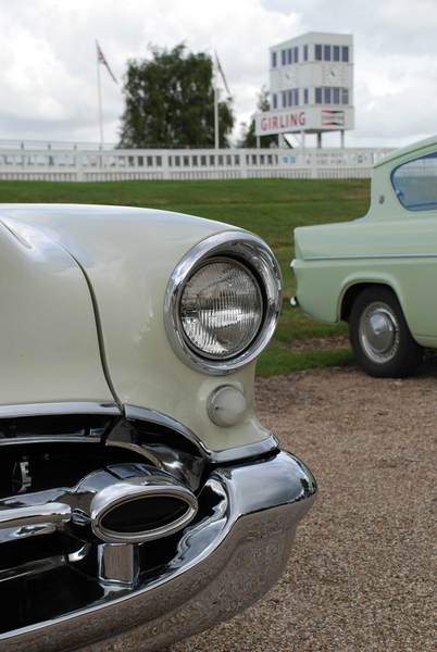 Breakfast August Pistonheads - The image showcases a vintage car scene, prominently featuring a classic white car with chrome accents carving an arc on a rocky path. The unique headlight design stands out, resembling a set of square eyes. The background is set against a clear sky, and a quaint white building with a clock tower can be seen in the far distance. The overall atmosphere suggests a peaceful, scenic location.