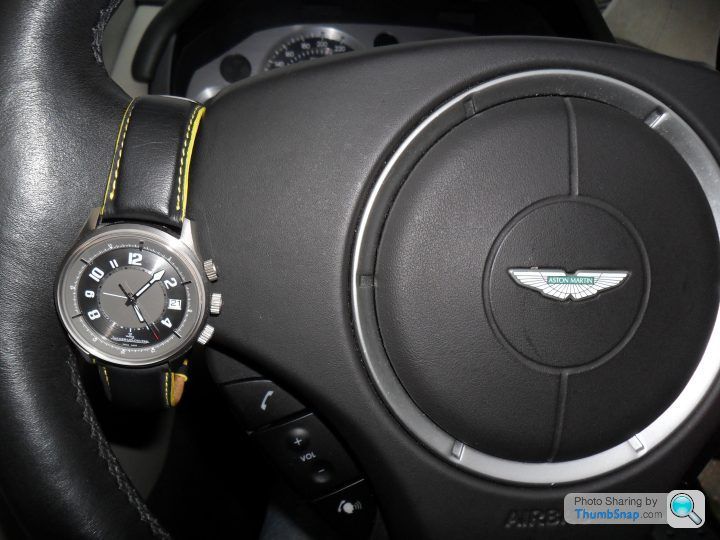 New V8V owner, let me introduce myself... - Page 1 - Aston Martin - PistonHeads - The image shows an indoor setting, focusing on the steering wheel of a car. The wheel is black with a metallic silver emblem in the center, below which there is a large analog clock face with black numerals and hands, encased within a gold-colored bezel. A yellow-green stripe is positioned at the 12 o'clock position on the steering wheel. The steering wheel is attached to a black dashboard that has various controls surrounding the central clock area. The overall aesthetic suggests a luxury car branding, possibly from Bentley, as indicated by the emblem.