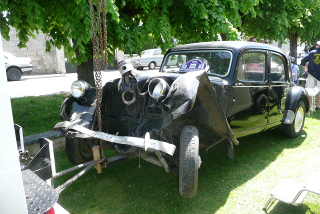 Spotted Commercials Classic Pistonheads - The image features an old-fashioned, dark-colored car parked among trees. The car's front hood is significantly damaged and holds no windscreen. A chain is hanging from the roof, and the car is located next to a parking meter. The tree provides shade, and there is a single chair visible toward the bottom right corner of the image. The paint on the rest of the car is showing signs of wear and rust, suggesting its vintage nature.