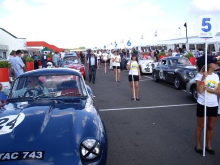 Silverstone Xks Pistonheads Historic - The image showcases an outdoor gathering around a range of classic cars. In the foreground, antique vehicles are on display, their polished surfaces reflecting the light, with numbers 24 and 743 prominently visible. Amidst the picture, several people are walking around, dressed in casual attire and a few are holding umbrellas. The setting appears to be a fair or exhibition, indicated by the presence of numbered markers among the vehicles. The ground is marked with white lines, possibly for designation or safety reasons. The open sky above and the clear visibility of the vehicles suggest a sunny day. Overall, the scene captures a dynamic and engaging atmosphere, likely centered around a car show or similar event.