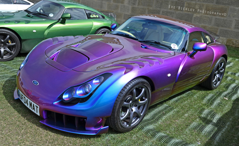 Chatsworth photos 2013 - Page 1 - TVR Events & Meetings - PistonHeads - In the image, there are two cars, one is a vibrant purple race car and the other is a flat green drag car. They are placed side by side on grass, positioned as if they are racing against each other. These cars are an example of the vivid colors and sleek designs associated with racing and drag culture. The image captures the excitement of mechanical sports and showcases the beauty of these high-performance machines.