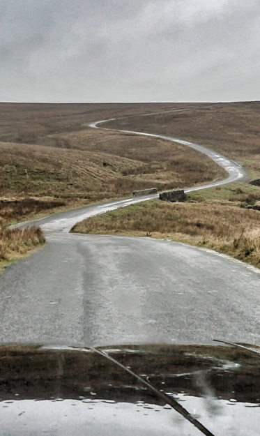 Kermit and co - the Pub2Pub TVR, and other steeds. - Page 3 - Readers' Cars - PistonHeads UK - The image is a photograph with an overlaid digital art effect, creating a layered visual. It shows a winding road in a rural landscape with rolling hills and verdant fields. The sky is overcast, casting a muted light on the scene. There's a vehicle visible at the bottom of the frame, suggesting the perspective of someone inside, looking out through the windshield. The style of the image merges realistic photography with an artistic alteration, possibly intended to evoke a sense of nostalgia or contemplation.