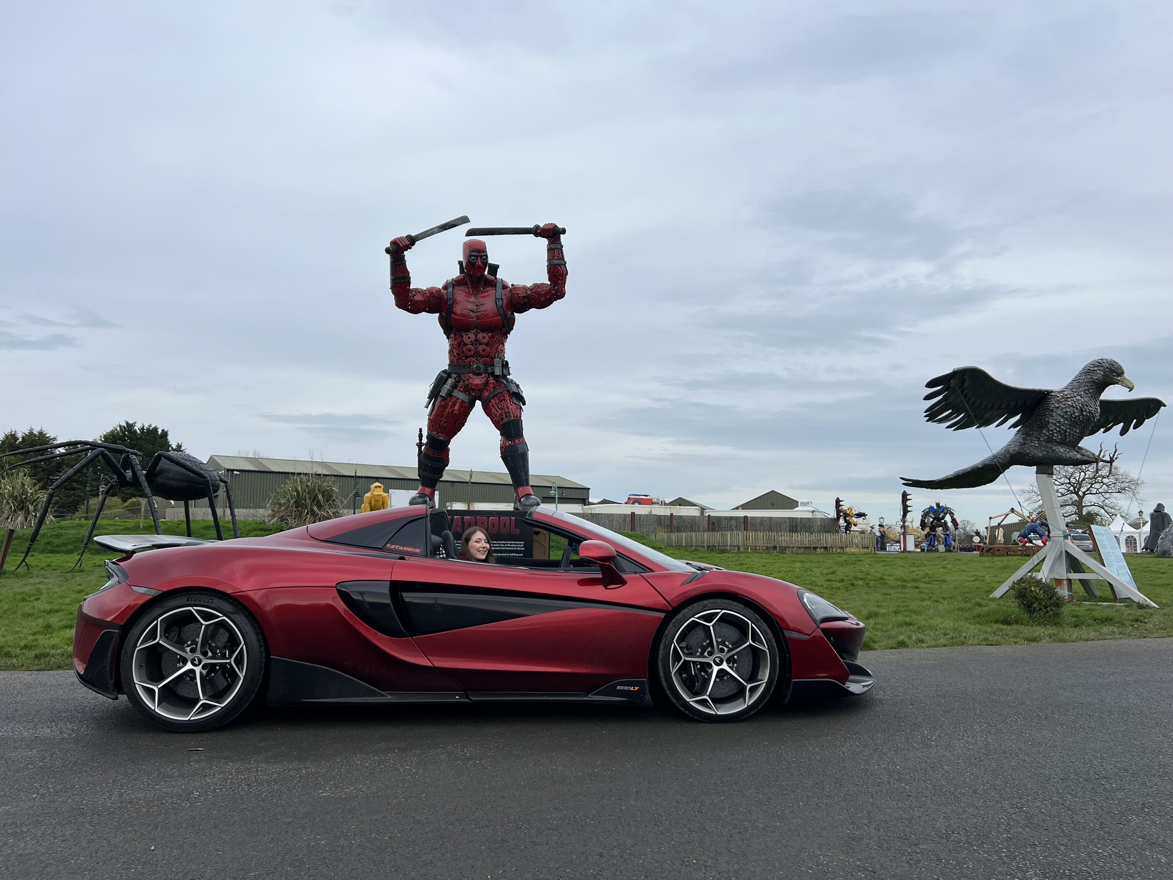 Pistonheads - The image captures a striking scene of a red sports car driving on the street. Intriguingly, a person dressed in a superhero costume is standing atop the car, giving an impression of levitation. The car itself is a stunning model, with its sleek body and distinctive features. It's parked on a wet road that reflects the surrounding environment. In the background, there are various objects including birds and a statue, adding depth to the image.