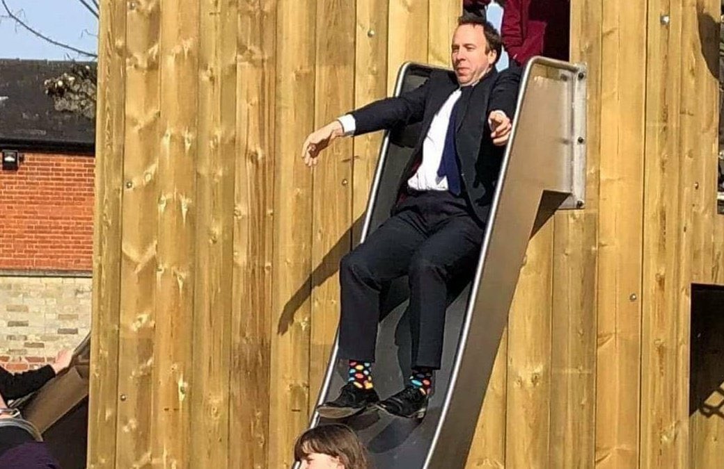 The Right Honourable Matt Hancock MP (Vol 1). - Page 5 - News, Politics & Economics - PistonHeads UK - The image depicts a man in a suit sitting on a large slide. He appears to be at the end of the slide, about to descend. The slide is located outdoors and there are people present who seem to be looking on. The setting includes a fence and some greenery, suggesting it might be an urban park or recreation area. The man's position suggests that he is either in the process of going down the slide or has just finished using it.