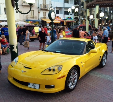 In Malta last week - Page 1 - Yank Motors - PistonHeads - The image captures a vibrant night scene on a street. The main focus of the image is a bright yellow Corvette sports car parked near the curb. The car is sleek and shiny, reflecting some of the street lights. There are several people standing around the car, perhaps admiring its design or engaging in conversation. The street is lined with shops and stores, their lighting contributing to the overall atmosphere of a bustling city nightlife. It's difficult to make out details about the people due to the lighting conditions and the focus on the sports car.