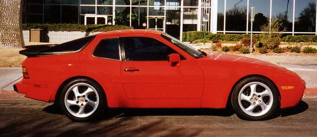 Which car best sums up the 1980s? - Page 7 - General Gassing - PistonHeads - The image shows a vibrant red sports car parked in a lot. The car's design is sleek and aerodynamic, indicative of a sports or racing model. It's parked in front of a modern building with large glass windows, suggesting an urban setting. The car is likely a Porsche model, judging by the distinctive emblem and design cues.