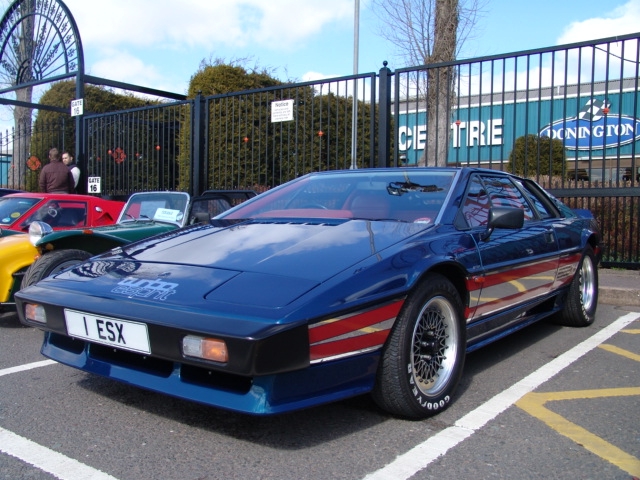 Espirt Guigaro celebration model  - Page 1 - Esprit - PistonHeads - The image shows a classic blue and red sports car parked in a parking lot. The car has a distinctive racy design, featuring angular lines, a large air intake on the engine bay, and a rear spoiler. There are a few people in the background, probably pedestrians. In the distance, there is a chain-link fence that encloses the parking area, and beyond it, a building is partially visible with a sign that reads 'AUTO CENTRE'. The sky is clear with a few clouds, suggesting good weather.